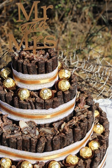 This wedding biltong cake has accents of gold and hessian made with 3 tiers for guest to enjoy. The biltong and droewors cake is custom made for a safari wedding with the addition of Ferrero Rocher chocolates to compliment the theme of the wedding and it adds a beautiful touch to the desert/snack table. MK Biltong Imagineer can custom design and make a biltong cake for you in any colour and size for your special occasion. Bush Themed Wedding, Boere Wedding Ideas, Biltong Cake, Bushveld Wedding Ideas, Biltong Cake Ideas For Men, Biltong Cake Ideas, Biltong Platter Ideas, Biltong Recipe South Africa, Bush Wedding South Africa