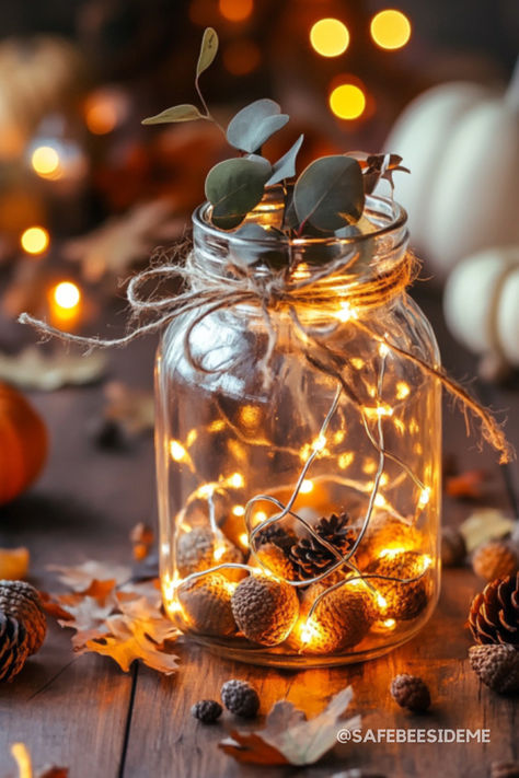 A mason jar centerpiece with warm fairy lights, acorns, pinecones, and eucalyptus sprigs tied with rustic twine, surrounded by autumn leaves and warm candlelight on a wooden table. *We may earn a small commission from your purchase. Mason Jar Fairy Lights Centerpiece, Jar Of Lights, Mason Jar Fairy Lights, Fairy Lights In A Jar, Mason Jar String Lights, Gallon Glass Jars, Indoor Photoshoot, Fall Mason Jars, Lighted Centerpieces