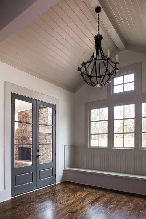 Gray cottage mudroom features a gray shiplap vaulted ceiling accented with an iron chandelier illuminating a gray beadboard window seat bench across from black glass paned doors. Beadboard Window, Chandelier Cottage, Gray Beadboard, Cottage Mudroom, Gray Cottage, Cottage Doors, Cottage Laundry Room, Gray Shiplap, Window Bench Seat