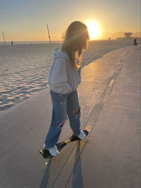 Skateboarding during sunset, at Venice Beach, California, woman on a Ripstick skateboard, wearing an oversized zip-up grey hoodie, baggy jeans with holes, and white tennis shoes, next to the sand and the ocean waves. Skateboard Beach, Ripstik Skateboard, Skateboard Beach Aesthetic, Beach Skater Aesthetic, Ripstick Skateboard Aesthetic, Beach Skateboard, Ripstick Skateboard, Venice Beach Aesthetic, Skateboard Photoshoot