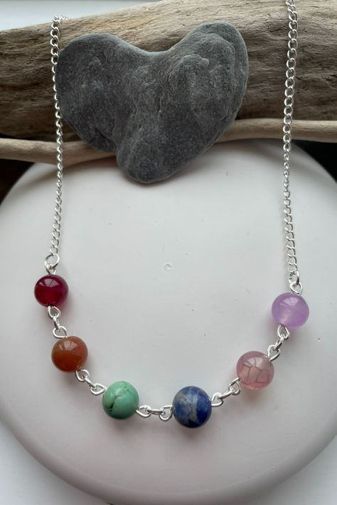 A silver plated chain with six gemstones in rainbow colours. The gems are: garnet, carnelian, howlite, sodalite, almandine and amethyst. The necklace rests on a white pebble and there are pieces of driftwood and a grey pebble at the top of the photo. Handmade Gemstone Necklaces, Silver Gemstone Beaded Necklaces For Festival, Silver Gemstone Bead Necklaces For Festivals, Silver Gemstone Beads Necklace Nature-inspired, Silver Nature-inspired Necklace With Gemstone Beads, Stone Necklace Diy, Silver Multi-stone Beaded Necklace, Semi Precious Stone Jewelry, Boho Jewellery