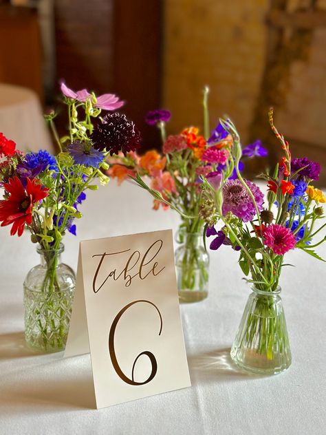 Three glasses vases filled with abundant natural flowers in jewel tones sit in a table clothed in white next to a sign which says Table 6. Simple Bright Wedding Centerpieces, Wedding Natural Decorations, Colorful Wedding Table Decorations, Bright Colorful Centerpieces, Bright Flower Centerpieces Wedding, Colorful Flowers Wedding Decoration, Simple Bright Wedding Flowers, Bud Vases Colorful Flowers, Colorful June Wedding Flowers