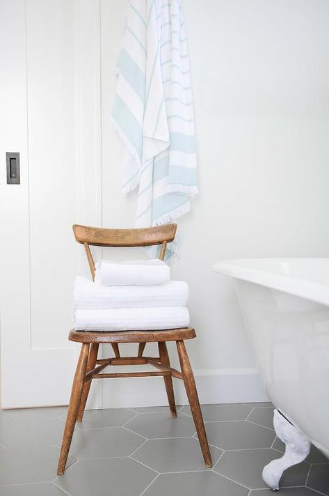 White and gray bathroom styled with a brown wooden vintage chair storing folded towels beside a white clawfoot tub atop large gray hexagon floor tiles. Bathroom Chair Ideas, Chair In Bathroom, White And Gray Bathroom, White Clawfoot Tub, Make Your House Look Expensive, Hexagon Floor Tiles, Folded Towels, Boho House Decor, Bathroom Chair