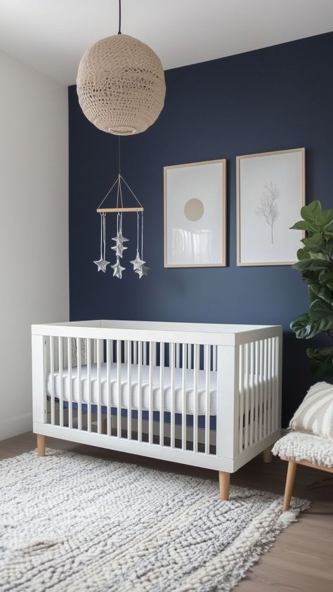 A moody nursery design with a white crib, a navy blue accent wall, minimalist framed art, a woven pendant light, and a mobile with silver stars. A plush neutral rug and a cozy chair with a striped pillow complete the look. Navy Blue And Beige Nursery, Modern Blue Nursery, Navy Blue And White Nursery, Navy And White Nursery, Boy Nursery Blue Accent Wall, Blue Nursery Paint Colors Sherwin Williams, Planetarium Nursery, Navy Blue Nursery Girl, Gender Neutral Blue Nursery