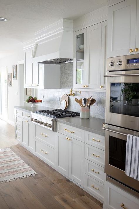 Polished brass hardware accents white kitchen cabinets topped with a gray quartz countertop holding a stainless steel gas integrated cooktop in front of gray marble subway backsplash tiles and beneath a white wood range hood. Kitchen Colors Paint, Grey Kitchen Ideas Modern Gray, Grey And White Kitchen Ideas, Grey Kitchen Ideas Modern, Grey Kitchen Ideas, Grey And White Kitchen, White Cabinets White Countertops, White Kitchen Ideas, Top Kitchen Cabinets