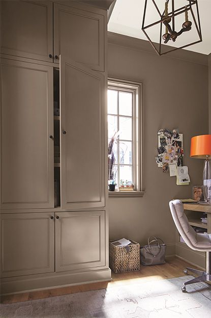 A taupe-painted home office with cupboard, beige chair, wooden desk, note board, white ceiling, and abstract chandelier. Dark Beige Kitchen Walls, Dellwood Sand Benjamin Moore, Best Dark Beige Paint Color, Furniture For Dark Walls, Rich Taupe Paint Color, Built In Bookcase Color Ideas, Falcon Brown Benjamin Moore, Taupe Walls And Trim, Taupe Wall Paint Colors