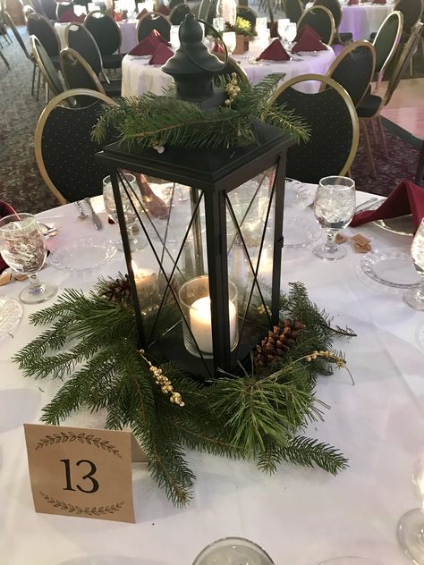 One of four centerpieces I made for my Michigan Winter Wedding! Candle, lantern, greenery, pine cone, gold accent. Follow me at __hannahhisey on instagram for more details on my winter wedding! Christmas Wedding Lantern Centerpieces, Winter Wedding Lanterns, Winter Wedding Lantern Centerpieces, December Wedding Decor Ideas, Pine Greenery Wedding, Winter Lantern Centerpieces, Winter Wedding With Christmas Trees, Pine Cone Wedding Centerpieces, Evergreen And Gold Wedding