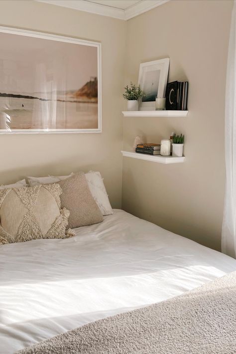 bedroom; white bedspread with tan accent pillows and throw blanket. beach photo with neutral colors above bed. shelving next to bed with books, plants, and candles. Bed Against Wall Nightstand, Small Room Ideas No Windows, Corner Wall Shelves Bedroom Small Spaces, Bedroom No Room For Nightstand, Small Bedroom Ideas Bed Against Wall, King Bed In Tiny Room, Bed Against Wall Decor, King Bed Small Space, Wall Space Ideas Bedroom