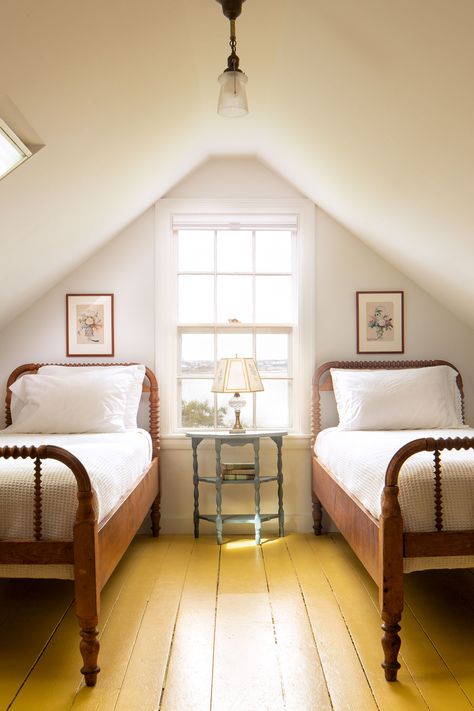 This restored Maine island home retains many of its original summer cottage details. Two twin spool beds frame a window with ocean views. Painted yellow floorboards accentuate the natural light that floods through a skylight in the sloping ceilings of the redone attic guest bedroom. Other antique details, such as a glass pendant light, light blue nightstand, and glass table lamp are fitting. New England Style Bedroom, Vintage Bedframe, Attic Guest Bedroom, Frame A Window, Attic Guest Room, Blue Nightstand, Spool Bed, Beds Frame, Stunning Bedrooms