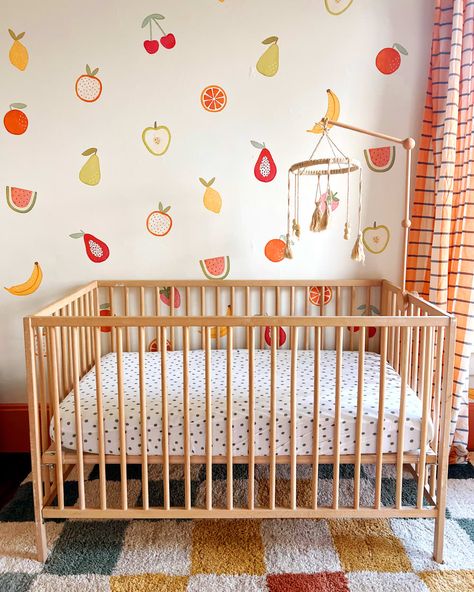 Macrame mobile above wood crib in nursery with fruit painted on wall. 70s Floral Nursery, Colorful Unisex Nursery, Nursery Fruit Theme, Painting Nursery Walls, Fruit Nursery Decor, Food Themed Nursery, Colorful Baby Girl Nursery, Painted Nursery Walls, Girl Nursery Colorful