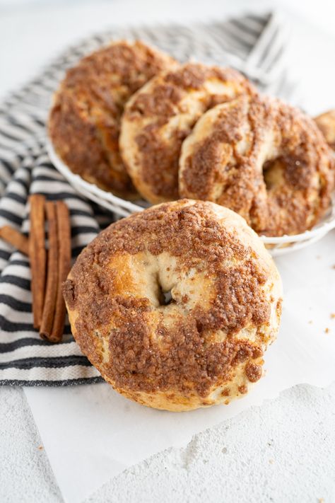 A bowl of cinnamon crunch bagels on the counter next to cinnamon sticks. Homemade Cinnamon Raisin Bagels, Sourdough Cinnamon Crunch Bagels, Cinnamon Streusel Bagels, Cinnamon Sugar Bagels, Cinnamon Bagel Recipe, Cinnamon Crunch Bagel Recipe, Cinnamon Crunch Bagel, Bagels Recipe Homemade, Cinnamon Bagel