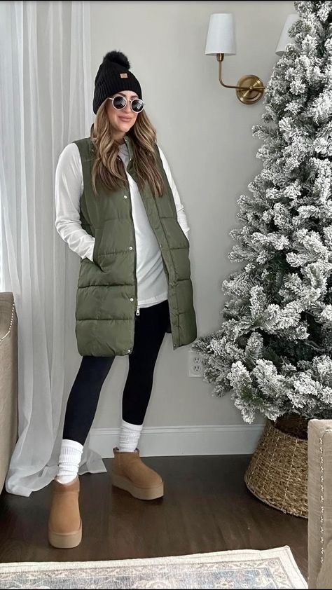 Woman in a holiday setting wearing an olive green long vest, white shirt, leggings, and tan boots, standing next to a snowy Christmas tree. White Puffy Vest Outfits For Women, Army Green Puffer Vest Outfit, Long Puffy Vest Outfits For Women, How To Style Vest Women, Olive Green Vest Outfits For Women, Sweatshirt Vest Outfit, Faux Leather Puffer Vest Outfit, Oversized Vest Outfits For Women, Short Vest Outfits For Women