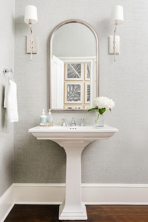 Between nickel sconces mounted to a wall covered in gray wallpaper, an arched beaded mirror hangs over a white pedestal sink finished with a polished nickel cross handle faucet. Powder Room Ideas With Wood Floors, Modern Bathroom Pedestal Sink, Mirror For Pedestal Sink, Pedestal Sink Mirror, Half Bathroom Pedestal Sink, Mirror Over Pedestal Sink, Staged Bathroom, Powder Room With Pedestal Sink, Pedastal Sink