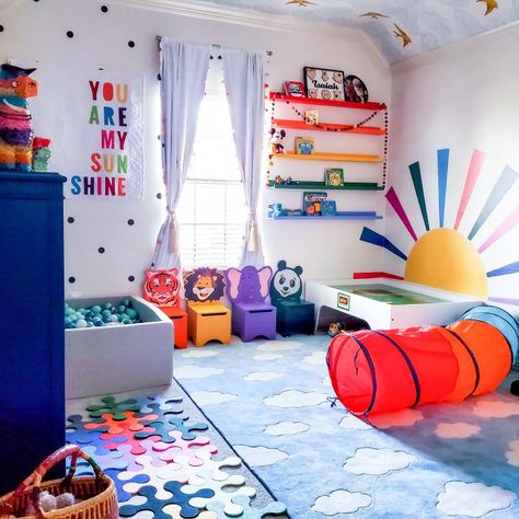 Bright blur rug with white fluffy clouds in center of room. Rainbow color shelves on a wall. Large horizon rising sun with rainbow colored beams. Large ball pit filled with an array of blue hued balls. Black and white polka dots on wall Bright Color Playroom, Playroom Ideas Colors, Nursery Decor Bright Colors, Bright Rainbow Bedroom, Playroom Bright Colors, Playroom Ideas Rainbow, Playroom Decor Colorful, Fun Colorful Nursery Ideas, Colorful Toy Room