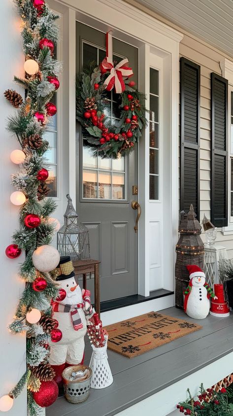 A cozy porch featuring a large red front door adorned with a festive green wreath. Lush garlands wrap around the doorframe, with twinkling white string lights and red bows. Exterior Xmas Decorations, Red And White Christmas Porch Ideas, Xmas Porch Decorating Ideas, Christmas Decor Ideas Red And White, Christmas Front Porch Decorating Ideas, Classic Christmas Decor Ideas, Door Garland Christmas, Red And Green Christmas Decor, Christmas Exterior