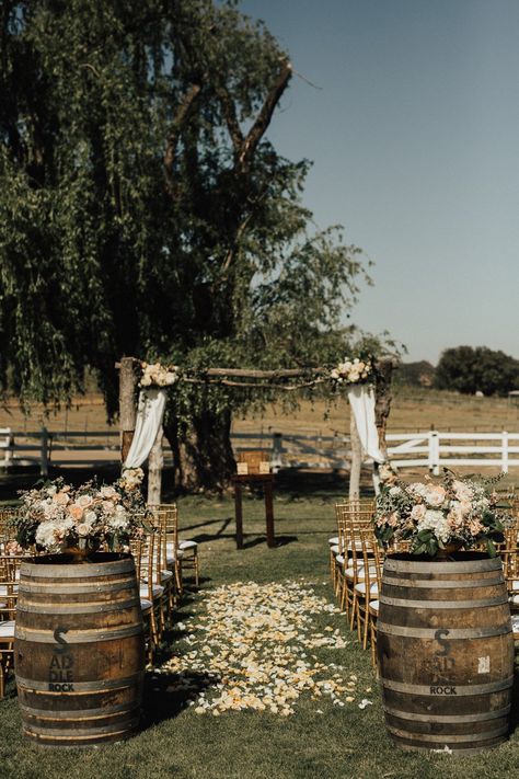 Two wine barrels with florals were at the base of the aisle, which was covered in flower petals. Outdoor Wedding Aisle Inspiration, Wine Barrel Ceremony Decor, Wine Barrel Wedding Aisle, Barrels At Wedding Ceremony, Wedding Arch With Barrels, Boho Ceremony Decor Aisle, Whiskey Barrel Wedding Decor Boho, Wooden Barrels Wedding, Whisky Barrels Wedding