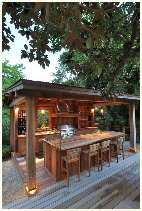 Rustic Outdoor Kitchen1 Outdoor wooden kitchen with bar seating, stainless steel appliances, and warm lighting under a canopy amidst lush greenery. | Sky Rye Design Outdoor Kitchen Modern Farmhouse, Diy Outdoor Kitchen On A Budget, Small Backyard Kitchen, Covered Grill Area, Garden Kitchen Ideas, Garden Kitchen Outdoor, Summer Kitchen Outdoor, Outside Kitchen Ideas, Cheap Outdoor Kitchen Ideas