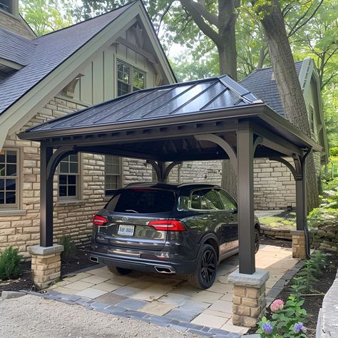 Elegant carport featuring a dark gray metal roof, beautifully complemented by tan stone accents and limestone piers. This open-sided structure shelters a black Volkswagen Tiguan, set against an old cottage-style house with black windows. The landscaping boasts lush greenery, colorful flowers, and two large decorative brown blocks enhancing the aesthetic.