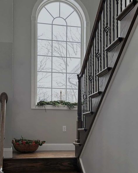 Dark wooden steps let down to a landing that features soft gray walls and a gorgeous arched window styled for the holidays with a garland and simple candle holder. The trim of the window is a classic white that matches the grilles. Staircase Wall Design Modern, Stairwell Window, Modern Stairway, Staircase Wall Design, Stair Bookshelf, Mirror Stairs, Staircase Window, Stairway Wall, Bookshelf Modern