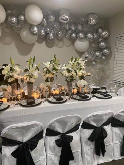 Glam up the dinner table for your family reunion with a charming white tablecloth and top it with a black runner. Line up tall cylindrical glass vases adorned with rhinestone wraps along the center and insert faux calla lily stems in them. Place white dinner plates, cutlery, and black linen napkins to perfect the setup while decorating the backdrop wall with white, silver latex, and orbz balloons. Also, cover the chairs with polyester white covers and black sashes for fabulous grandeur. Silver Dinner Party, Black Linen Napkins, Event Decor Ideas, Bday Dinner, Backdrop Wall, White Tablecloth, Black Runners, White Dinner, White Dinner Plates