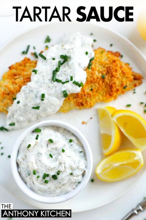 An overhead shot of a piece of fish covered in tartar sauce.  Also on the white plate are lemon wedges and a small bowl of tartar sauce.