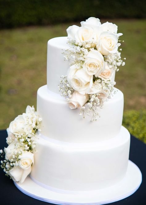 A White and beautiful wedding cake. The white wedding cake is decorated with white roses. The white wedding cake is made into three tiers. White Roses Decoration Wedding, Wedding Cakes With Gypsophilia, Wedding Cake 3 Tier Flowers, White Wedding Cake With White Flowers, White Roses Wedding Cake, White Roses On Wedding Cake, Three Tier White Wedding Cake, Wedding Cake With Baby Breath Flowers, 3 Tier White Wedding Cake With Flowers