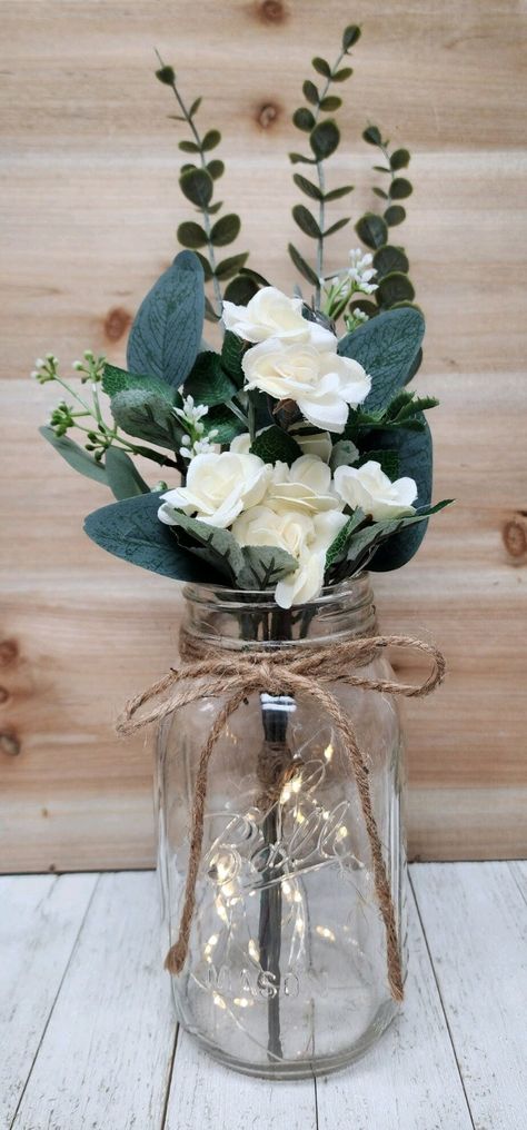 These beautiful faux mini-rose quart size mason jar centerpieces are a Glassy Gal original design. Each centerpiece includes one set of warm white LED fairy lights, a jute bow, a deluxe faux greenery arrangement with mini roses. You can choose your mini rose color at checkout.  These centerpieces are very popular for weddings, baby showers, parties, corporate events, holiday events, and any other event that requires table centerpieces. They also look great in any room of your house or office. To Boho Mason Jar Wedding Centerpieces, Mason Jar Centerpieces Table Decorations, Simple Centerpieces Wedding, Centerpieces Without Flowers, Wedding Shower Centerpieces, Jar Centerpiece Wedding, Barn Wedding Centerpieces, Mason Jar Flower Arrangements, Wedding Ceremony Readings