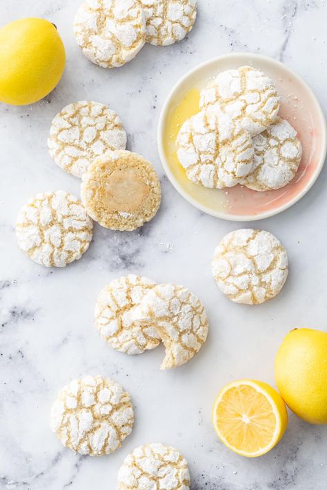 Overhead, scattered Chewy Lemon Almond Crinkle Cookies on a marble surface with a few fresh lemons and three cookies on a small ceramic plate. Lemon Turmeric Crinkle Cookies Nyt, Almond Egg White Cookies, Lemon Powder Cookies, Lemon Amaretti Cookies, Lemon Turmeric Crinkle Cookies, Lemon Tumeric Cookies, Lemon Curd Shortbread Cookies, Meyer Lemon Cookies, Recipes With Almond Extract