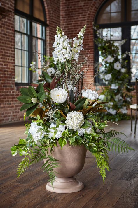 Set within a low-profile pair of taupe stone planters, the classic pairing of crisp white and lush green foliage, allow these stems, gradually building in height and texture, to capture visual interest and make an immediate statement! Blossoming magnolia, Chantilly-white Snowflake snapdragons, and creamy open-faced peonies pop against the lush greenery of Dixie Wood fern, Southern Shield fern, clematis, Golden pothos, and silver inch plant, while dried willow and limonium branch upwards, creating organic movement and delicate layers. The Beaumonts are simply perfect to anchor the altar space, can be elevated above a ground arch, begin the aisle way, or placed on either side of a table, bar or seating chart. The possibilities of these neutral and naturally elegant pieces are endless! Gorgeo Cream And Green Flower Arrangements, Foliage Centerpiece Wedding, Brown And White Flowers, Green And White Floral Arrangements, Magnolia Gardens, White Floral Arrangements, White Flower Arrangements, Greenery Arrangements, Large Floral Arrangements