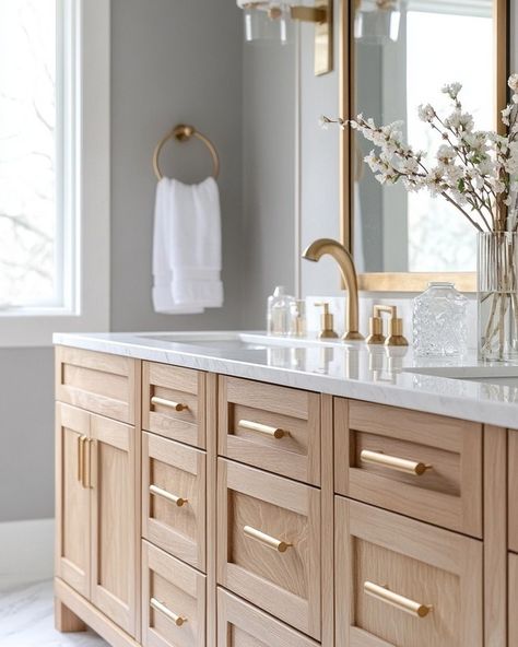 When it comes to creating a space that feels luxurious yet grounded, I love mixing natural tones and textures with sleek, modern fixtures. The oak cabinetry paired with the marble countertops and gold accents here creates that perfect balance of warmth and elegance, don’t you think? I’ve been obsessed with how the natural wood grains pop against the soft grey walls. And who knew a simple touch like fresh florals could transform the entire room? ⁣ ⁣ I’ve found that bringing in natural material... Light Wood And Gold Bathroom, Natural Oak Bathroom Vanity, Wood Vanity Gold Hardware, Natural Wood Bathroom Ideas, White Oak Kitchen Cabinets Gold Hardware, Light Wood Bathroom Cabinets, Natural Wood Bathroom Cabinets, Grey And Wood Bathroom Ideas, White Oak Bathroom Cabinets