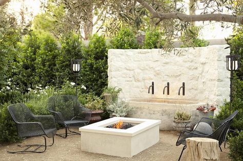 Patio seating area features a white concrete fire pit flanked by metal lounge chairs lit by torches positioned on either side of a curved white stone water fountain. Outdoor Fireplace With Fountain, White Pergola Ideas, Courtyard Fire Pit, Moroccan Backyard, Landscape Renovation, Outdoor Amenities, House California, White Pergola, Patio Seating Area