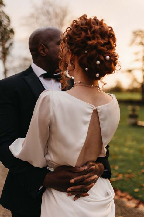 Groom in black tux with bowtie embracing bride in open back detail satin wedding dress with pearl hair accessories and pearl necklace Curly Updo With Pearls, Boho Chic Hairstyles Wedding, Curly Hair Pearls, Curly Updos For Wedding, Pearl Wedding Hairstyles, Bridal Updo Pearls, Wedding Updo Pearls, Shoulder Length Curly Wedding Hair, Pearl In Hair Hairstyles