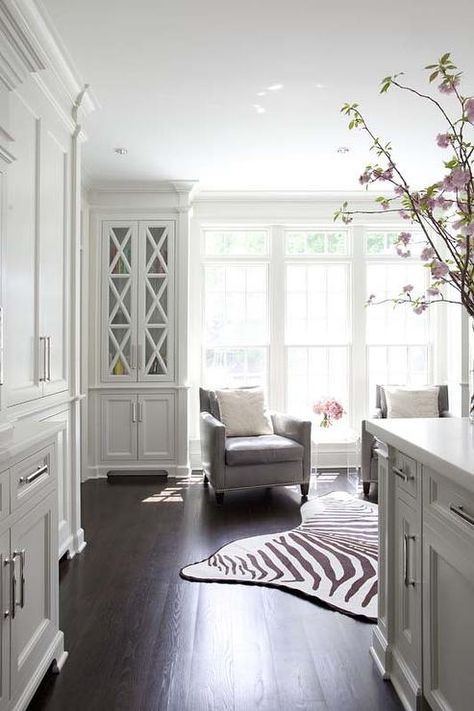 KItchen sitting room is filled with a pair of gray accent chairs flanking an acrylic accent table atop a zebra cowhide rug placed in front of windows flanked by built in bookcases with glass doors. White Sitting Room, Chairs In Kitchen, Sitting Area Design, Kitchen Sitting Room, Cabinet With Glass Doors, Grey Accent Chair, Lucite Table, Bookcase With Glass Doors, Built In Cabinet