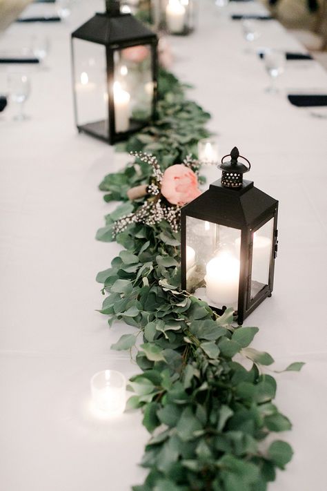 Black lanterns for winter wedding centerpieces and greenery table runner Black And Greenery Wedding Centerpieces, Lantern Centerpieces Long Table, Wedding Table Decor Lanterns, Black Lanterns Wedding Centerpiece, Winter Wedding Centerpieces Rectangle Table, Wedding Table Decorations With Lanterns, Black White And Greenery Wedding Centerpieces, Lantern Centerpieces With Greenery, Boho Lantern Centerpieces