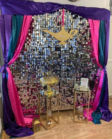 A silver sequin Backdrop draped in purple, Teal and pink material to give an arabian nights vibe. A hanging gold genie lamp with a name hangs in the middle and there are 2 gold plinths with a cake stand in front of the Backdrop. Arabian Nights Photo Booth, Arabian Nights Wedding Theme Decor, Aladdin Prom Theme, Arabian Nights Backdrop, Arabic Party Decoration, Arabian Night Decoration, Arabian Nights Decor, Arabian Night Decor, 1001 Nights Party