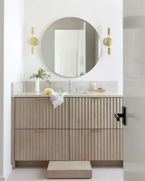 a greige fluted vanity with gold handles, a stone countertop, a round mirror, gold sconces and some lovely decor Fluted Bathroom Door, Greige Bathroom Vanity, L Shaped Bathroom Vanity, Fluted Bathroom Vanity, Grey Terrazzo Floor, Greige Bathroom, Tiny Home Bathrooms, L Shaped Bathroom, Fluted Vanity