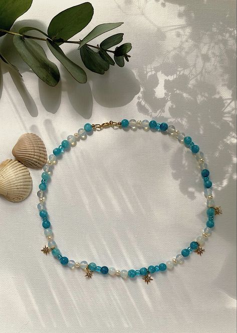 Handmade necklace, natural blue stone beads, opalite beads, blue glass beads, small freshwater pearl, gold pated cubic zircornia star charms, stars, blue ocean color, blue aesthetic, seacore, beach core style, sea inspired, 45cm necklace, beaded necklace. Displayed on a white background with 2 shells in the right side of necklace and green plant on the top. Sea Necklace Aesthetic, Color Blue Aesthetic, Beaded Necklace Aesthetic, Jewellery Creative, 45cm Necklace, Beach Core, Pretty Stars, Aesthetic Jewellery, Aesthetic Creative