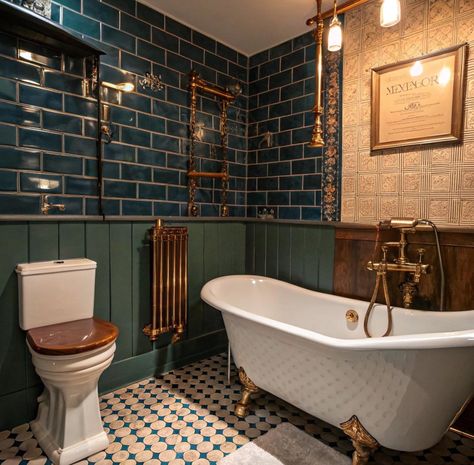 A vintage-style bathroom with deep green subway-tiled walls, dark green wainscoting, and brass fixtures. A freestanding white clawfoot tub with gold feet sits on patterned floor tiles. Above the tub, there’s a brass shower fixture and towel warmer. The room includes a framed wall piece and unique lighting, adding warmth to the classic decor. Art Deco Shower Room, Vintage Victorian Bathroom, 1930 Bathroom, Victorian Bathroom Vintage, Victorian Bathroom Ideas, Green Bathrooms, Green Subway Tile, Paneled Walls, Patterned Tile