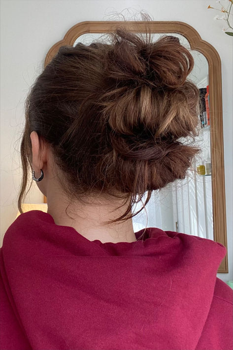 A hairstyle featuring three messy buns stacked together at the back of the head, creating an effortless and textured updo. The hair is soft brown with natural highlights, and a few loose strands frame the neck and sides. She is wearing a maroon hoodie, and the reflection in a wooden-framed mirror shows a bright, minimalist room with a cozy and natural feel. Medium Length Hairstyles Updo, Shoulder Length Hairstyles For School, Up Hairstyles For School, Medium Length Hairstyles For School, Hair Inspo Updo, Winter Hairstyles Medium, Hairstyles Updo Casual, Curly Hair Easy Hairstyles, Cute Hair For School