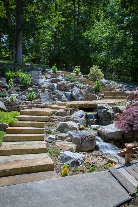 Modern Design Aquascaping did a tear-out & re-installation of a pondless waterfall in Lenoir City, TN. The feature which was done by Modern Design Aquascaping, includes large mountain boulders,cedar driftwood accents, LED under-water lighting, flagstone walkway-steps-and bridge, and a wooden bridge. Cascade Waterfall Garden, Backyard Creek Landscaping, Mountain Garden Ideas, Pondless Waterfall, Flagstone Walkway, Fountains Backyard, Sloped Backyard, Natural Pond, Pond Landscaping