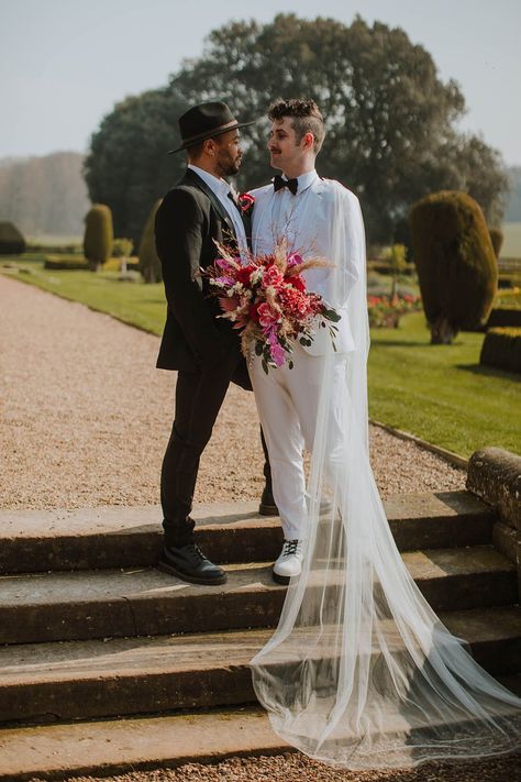 Groom in a black suit and fedora hat standing with his partner at Prestwold Hall Barns in a white groom suit and cape holding a vibrant red bouquet Wedding Suit With Veil, Mens Wedding Suit With Cape, Unconventional Wedding Suit, Wedding Suit With Cape, Celestial Wedding Suit, Enby Wedding Outfit, Fancy Wedding Suit, Enby Wedding, Gay Wedding Outfits