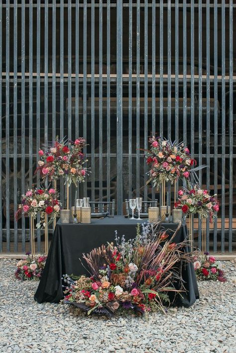 A wedding day sweetheart table is pictured. The table has a black tablecloth and black place settings with clear glasses. It is surrounded by floral arrangements that have red, pink, purple, and peach colored flowers along with dried flowers in assorted fall colors. Black Tablecloth Wedding, Burgundy Tux, Grunge Wedding, Gold Table Decor, Traditional Gown, Dark Elements, Table Floral Arrangements, Black Tablecloth, Sweetheart Table Wedding