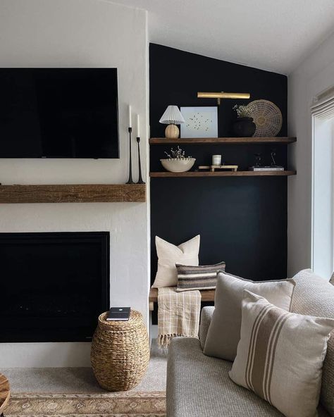 This neutral and black living room features a TV mounted over a warm wood mantle and a black accent wall fitted with warm wood floating shelves adorned with modern accents. A brass portrait light fixture mounted over the selves brings life to this corner. Large windows bathe the room with bright natural light and a bench layered with accent pillows rests below the shelving. Floating Shelf By Fireplace, Fireplace Shelves Floating, Floating Shelves Lighting, Moody Fireplace Wall, Neutral And Black Living Room, Styling Fireplace, Black Accent Wall Living Room, Shelves Lighting, Black Accent Wall