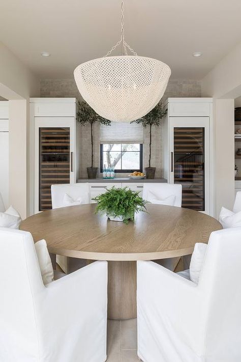 A white basket chandelier illuminates white slipcovered dining chairs placed around a round wood pedestal dining table. Pure Salt Interiors Dining Room, Salt Interiors, Modern Dining Room Ideas, Pure Salt Interiors, Transitional Dining Room, Transitional Dining, Coastal Dining, Modern Colonial, Pure Salt