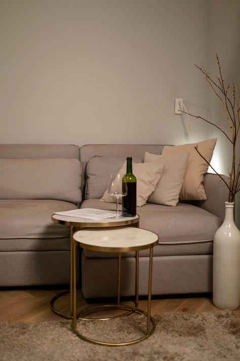 A cozy light gray sofa invites relaxation, featuring soft cushions that enhance comfort. The fabric showcases a subtle texture, perfectly complementing the warm-toned wooden floor beneath. Nearby, a stylish set of nested tables with gold metal frames support a bottle of wine and a delicate glass, adding a touch of elegance. Decorative elements, including patterned throw pillows and a slender vase with branches, create a harmonious atmosphere, making this setup an ideal space for unwinding and enjoying a moment of tranquility. Light Gray Sofa, Sofa Comfy, Light Gray Sofas, Vase With Branches, Marble Top Side Table, Classy Living Room, Designer Lamp, Modern Living Room Interior, Luxury Living Room Design