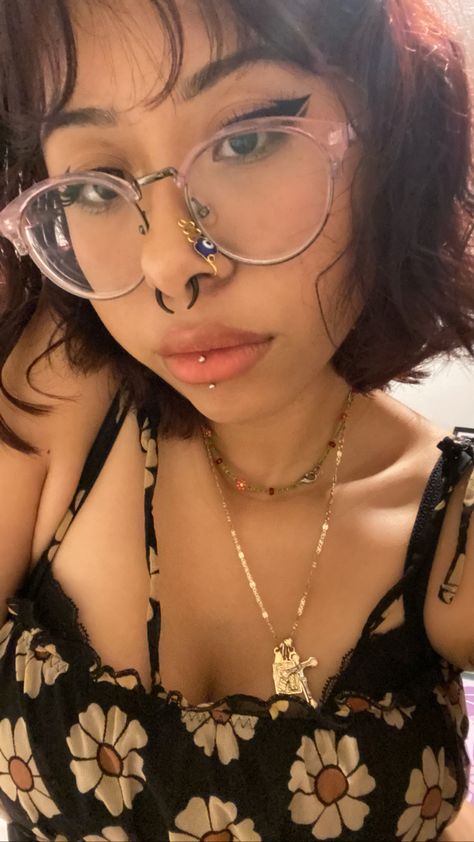 Female with short dark red hair. Wearing round purple glasses. Has a stretched 8 gauge septum with black horseshoe jewelry. Wearing an evil eye gold nose cuff. Has a vertical labret piercing with titanium jewelry. Is wearing a yellow flower and black spaghetti strap top. Has a gold necklace with a cross, and a shorter, seed beed flower necklace. Vertical Labret Piercing Jewelry, Central Labret Piercing, Vertical Labret Piercing Aesthetic, Vertical Labret Jewelry, Vertical Piercing, Central Labret, Septum Stretching, Cosmetic Aesthetic, Micro Bangs