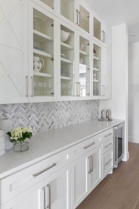 Stacked white butler's pantry cabinets accented with x-trim moldings flank stacked glass front cabinets located above a white and gray marble herringbone backsplash. Pantry Backsplash, White Herringbone Tile Backsplash, White Herringbone Tile, Herringbone Tile Backsplash, Marble Herringbone, White Marble Kitchen, White Kitchen Backsplash, Built In Pantry, Herringbone Backsplash