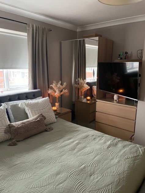 A side photograph of a bedroom showing half of the bed, bedside table, back wall and mirrored wardrobe with a reflection of the beside table - on which stands cream coloured pampas grass in a vase, with a black lit candle reflecting. The colours of the bedroom include Taupe walls, a sage green duvet cover featuring 2 white square pillows and 1 beige rectangular pillow. The image feels calming and relaxed. Sage Bedspread, Cream And Sage Bedroom, Sage Green Bedding, Sage Bedding, Sage Bedroom, Taupe Walls, Natural Bedroom, Beige Curtains, Calming Bedroom