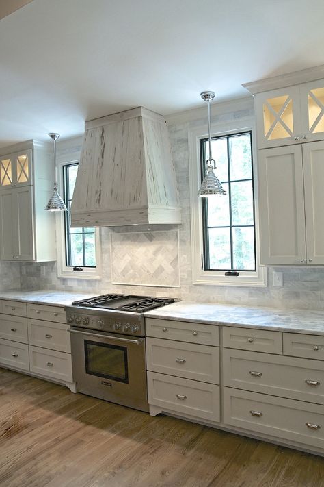 Grey kitchen. Stunning transitional kitchen is equipped with a rustic pecky cypress hood fixed between windows framed by gray marble backsplash tiles illuminated by industrial pendants. The hood is located above herringbone marble cooktop tiles accenting a Thermador range flanked by white drawers with polished nickel cup pulls topped with gray and white marble countertops positioned beneath white upper cabinets finished with x front lighted display cabinets. According to the designer, the ca... Kitchen Cabinets Grey And White, Kitchen Window Design, Kitchen Cabinet Trends, Hood Design, Kitchen Hood, White Shaker Cabinets, Herringbone Backsplash, Kitchen Hoods, Classic Kitchen