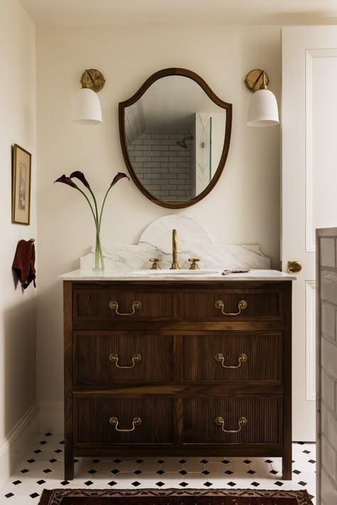 This elegant bathroom features a vintage-inspired wooden vanity with ribbed detailing and brass hardware, topped with a luxurious white marble countertop, complemented by a round, antique-style mirror, soft white wall sconces, and a neutral cream-colored palette, creating a warm and inviting ambiance, while the checkered black-and-white floor tiles add a touch of modern charm. Dark Wood Vanity Bathroom, Mcgee And Co Bathroom, Black And White Marble Bathroom, Vessel Sink Ideas, Half Bath Vanity, Dresser Vanity Bathroom, Wooden Bathroom Vanity, Antique Bathroom Vanity, Black And White Tiles Bathroom